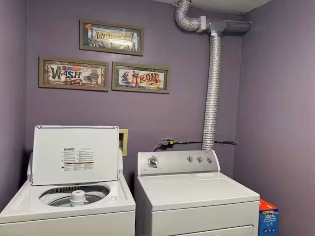 Un cuarto de lavado con dos lavadoras blancas, un conducto de ventilación y dos letreros enmarcados en la pared.