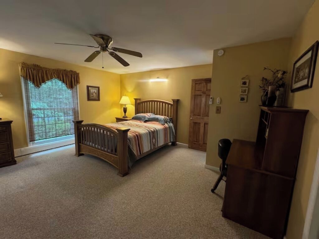 Un dormitorio con cama, ventilador de techo, ventana con cortinas, escritorio y silla.