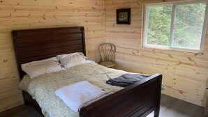 A wooden cabin bedroom with a bed, a chair, and a window. - “The Summit Cabin” <br>An Off the Grid Hideaway (Barnardsville)