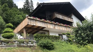 Ein zweistöckiges Holzhaus mit einem Balkon, umgeben von üppiger Vegetation und einer Steinmauer.
