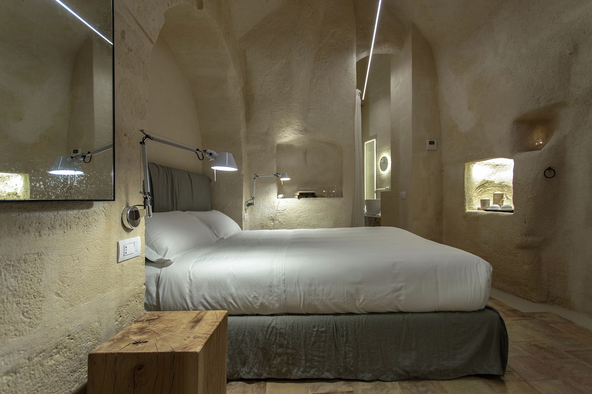 A bedroom with a large bed, a wooden nightstand, a mirror, and a small window.