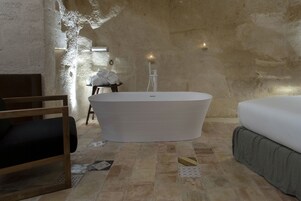 A room with a large bathtub, a wooden chair, a bed, and a small table with towels.