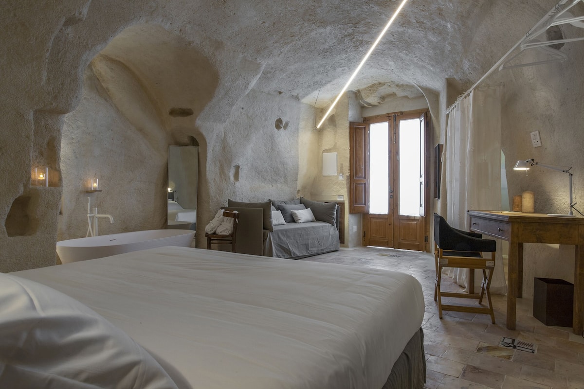 A bedroom with a large bed, a desk, a chair, a bathtub, and a door leading to another room.