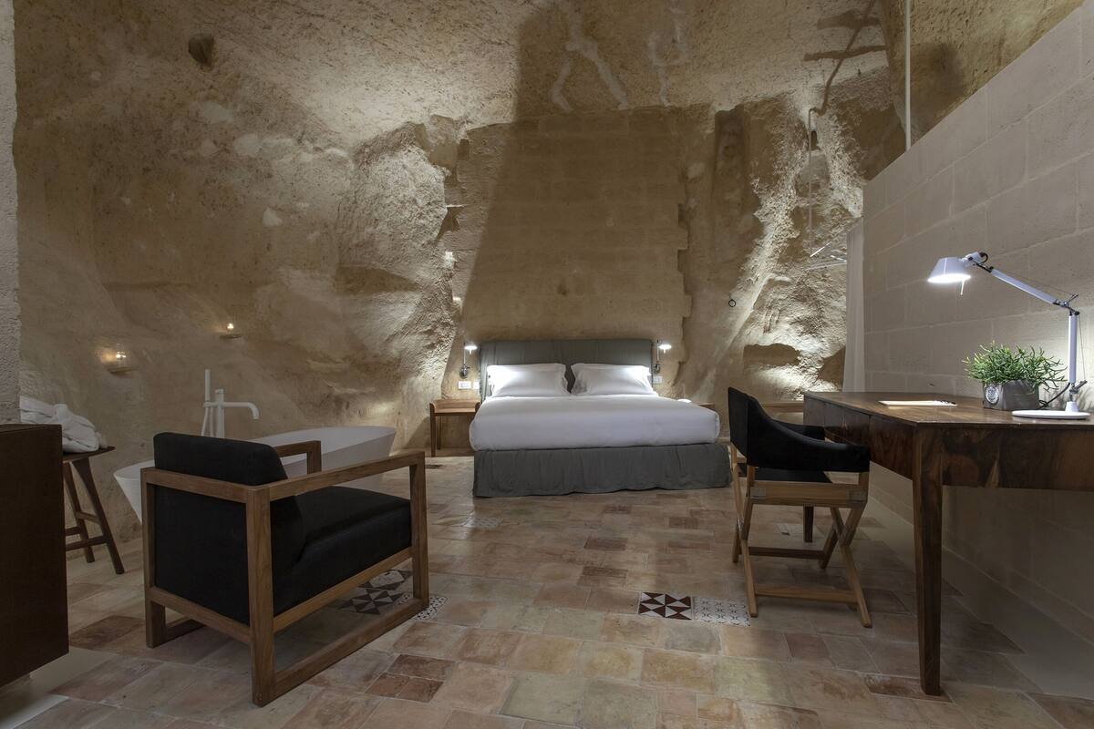 A bedroom with a stone wall, a bed, a desk, a chair, a bedside table, and a lamp.