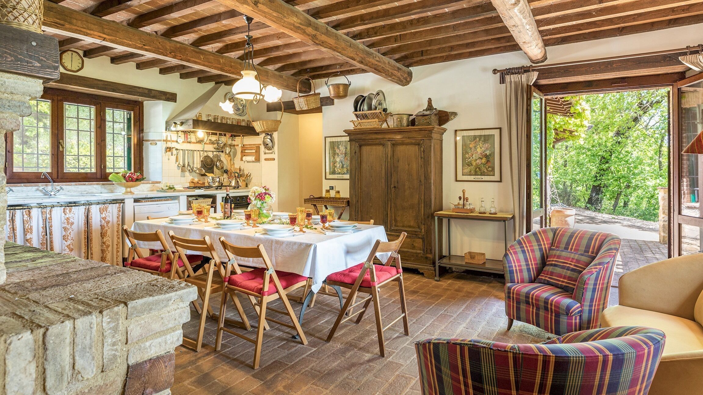 Un comedor rústico con una mesa de madera dispuesta para cuatro personas, rodeada de sillas de madera con cojines rojos. Se ve un aparador de madera grande y un sillón colorido al fondo.