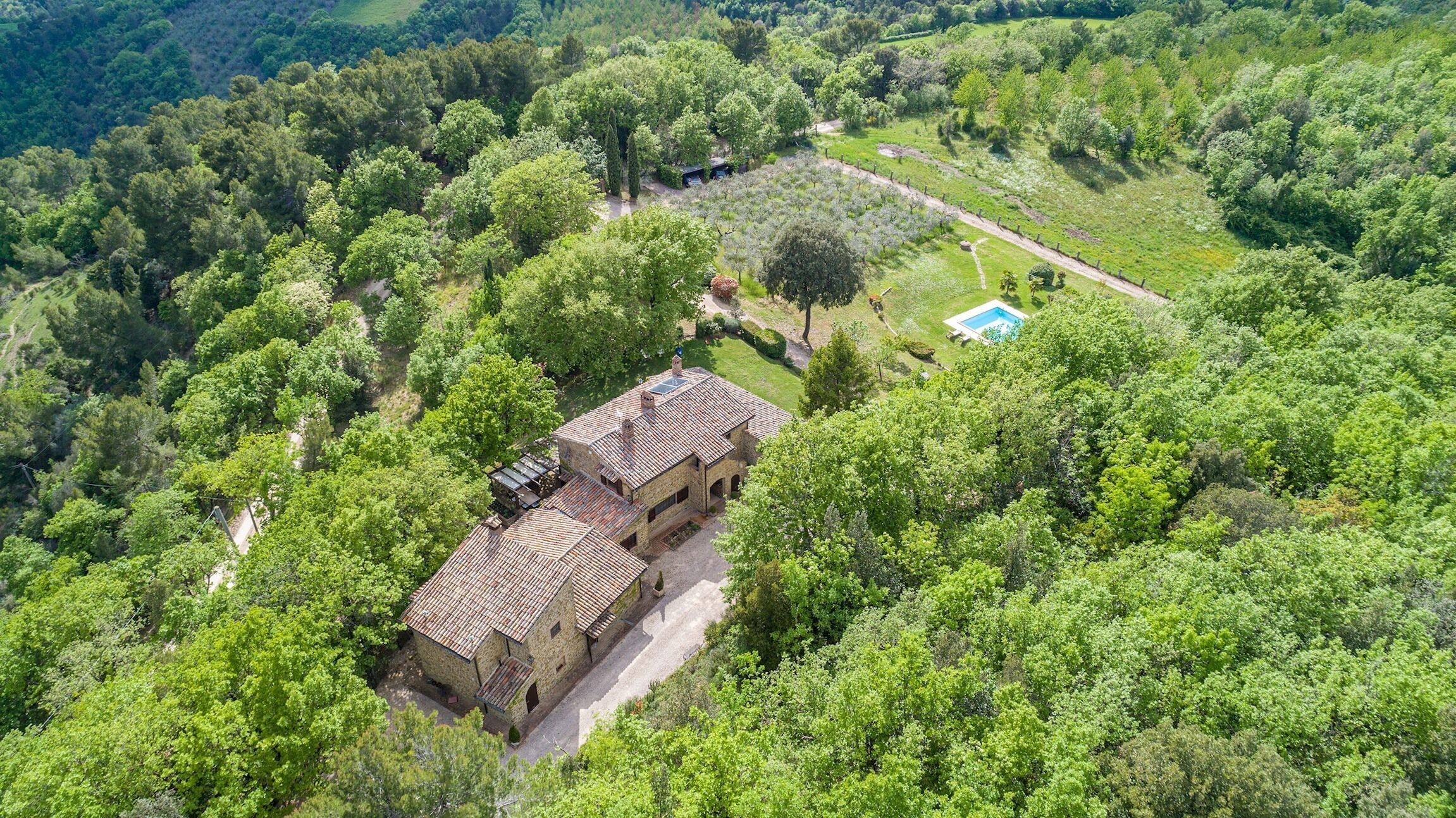 An aerial view of a stone house surrounded by dense greenery, with a visible swimming pool and a clear path leading to the house.