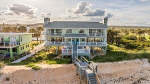 A two-story beachfront house with multiple balconies, a covered porch, and a sandy beach in the foreground. - Tipsy Turtle Direct Oceanfront Sunrise (Palm Coast)