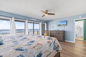 A bedroom with a large bed, a ceiling fan, a dresser, and a view of the ocean through large windows.