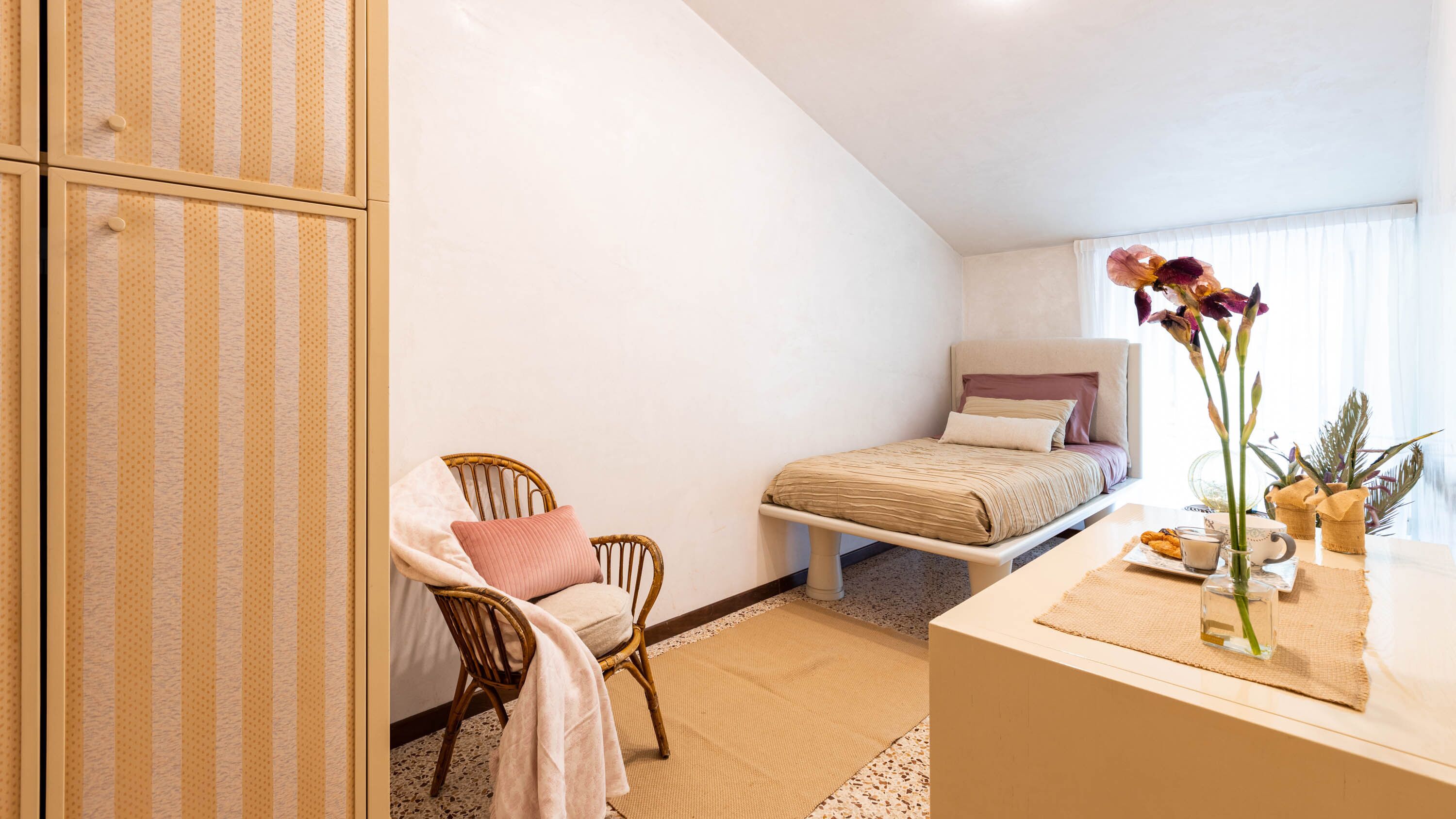 Ein Schlafzimmer mit einem Bett, einem Sessel, einem Nachttisch mit Blumenvase und einem Kleiderschrank.