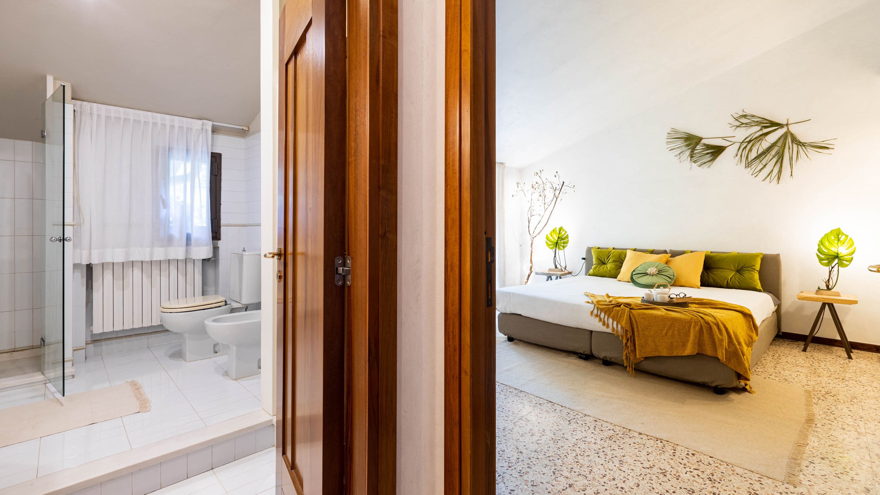Une salle de bain moderne équipée de toilettes, d’un bidet et d’un espace douche. La chambre comprend un lit, des tables de chevet et des plantes décoratives.