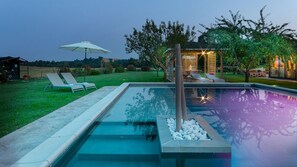Een zwembad met traptreden, ligbedden en een pergola met verlichting.