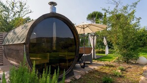 Eine große, zylindrische Struktur mit reflektierender Oberfläche, möglicherweise eine Sauna oder ein Whirlpool, umgeben von einem Garten mit einer Pergola und Sitzgelegenheiten.