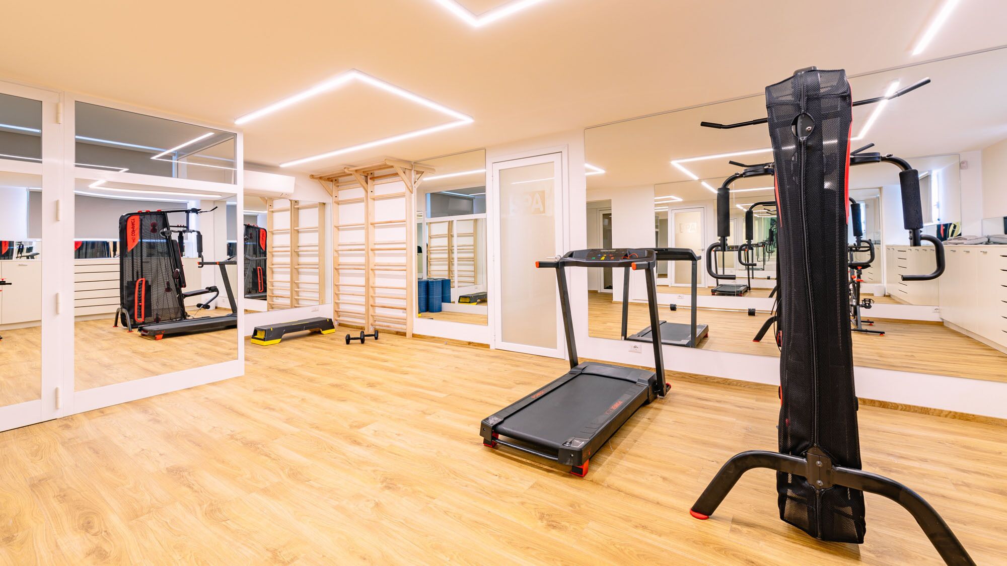 A modern gym with a wooden floor, various exercise equipment, and a large mirror reflecting the space.