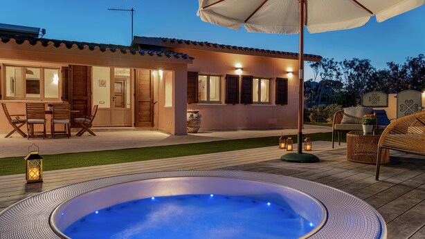 Un espace extérieur accueillant, avec un patio, un jacuzzi, des meubles en bois et une maison bien éclairée, dotée d’un toit en tuiles.