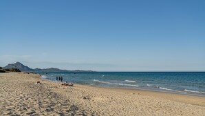Une vaste plage de sable fin, avec des vagues douces, un ciel dégagé et des montagnes à l’horizon.