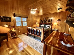 Une chambre dans une cabane en bois, comprenant un lit, un bureau, une chaise, un ventilateur de plafond et une fenêtre.