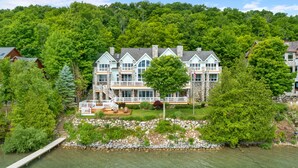 A large, multi-story house with a stone facade, multiple balconies, and a dock extending into a lake.
