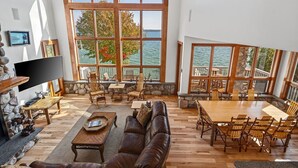 A spacious living area with a large wooden table and comfortable seating, overlooking a body of water through large windows.