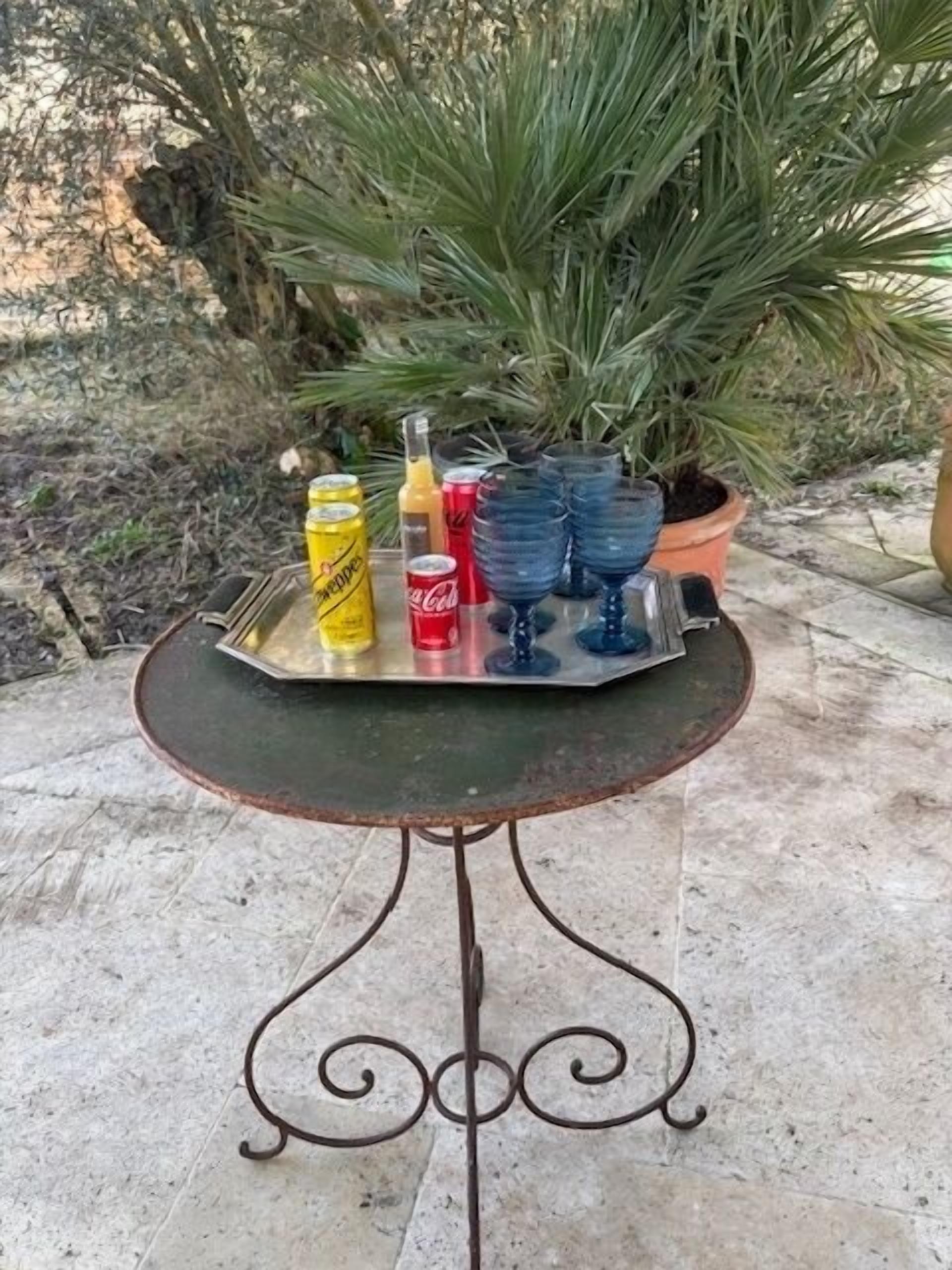 Ein Metalltisch mit einer Ablage, auf der sich eine Dose gelbes Getränk, eine Orangensaftflasche, eine rote Cola-Dose und mehrere blaue Gläser befinden.