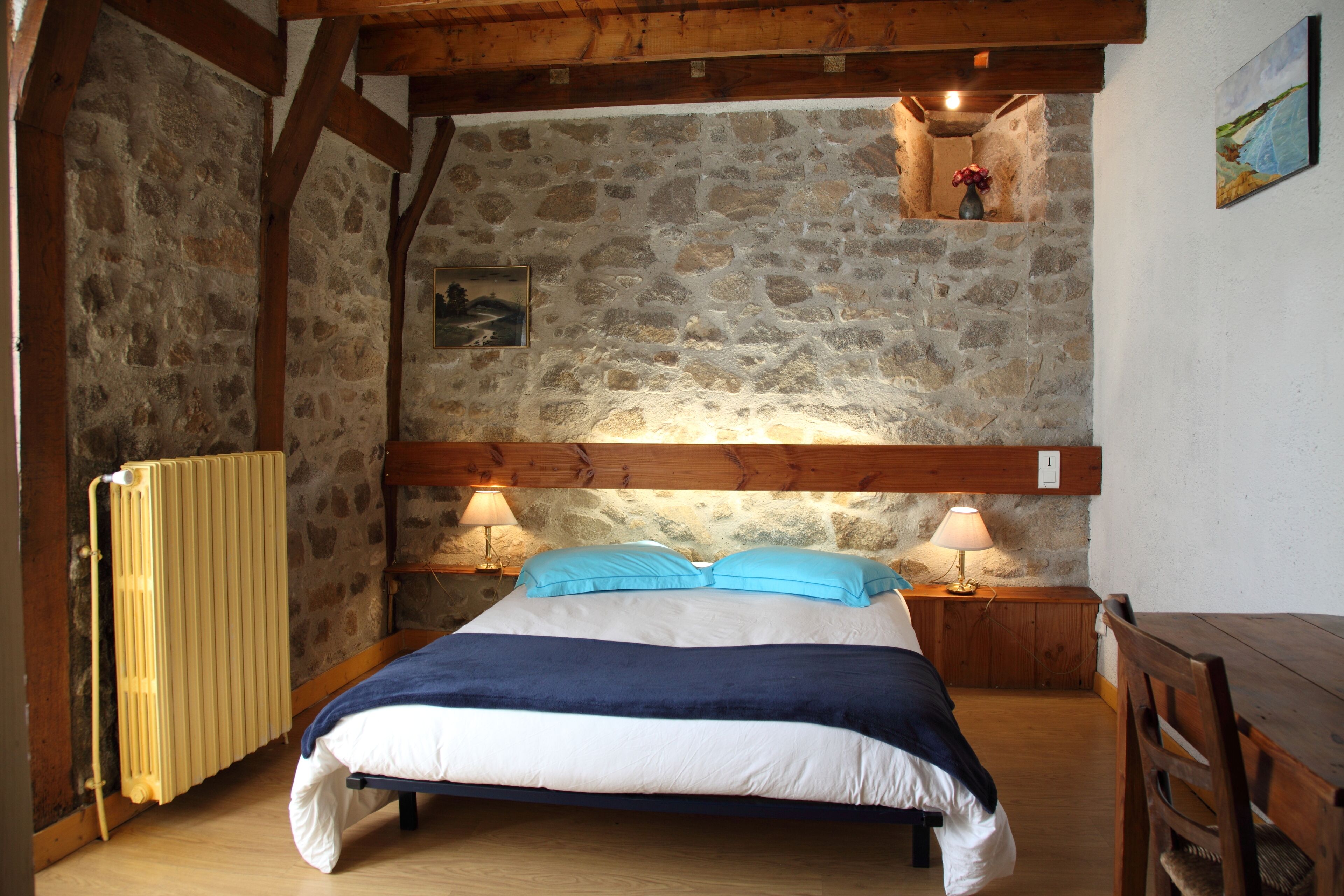 A bedroom with a stone wall, wooden beams, a bed with blue and white bedding, two bedside lamps, and a wooden table.