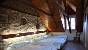 Een kamer met drie bedden tegen een stenen muur, houten balken in het plafond en een raam dat voor natuurlijk licht zorgt.