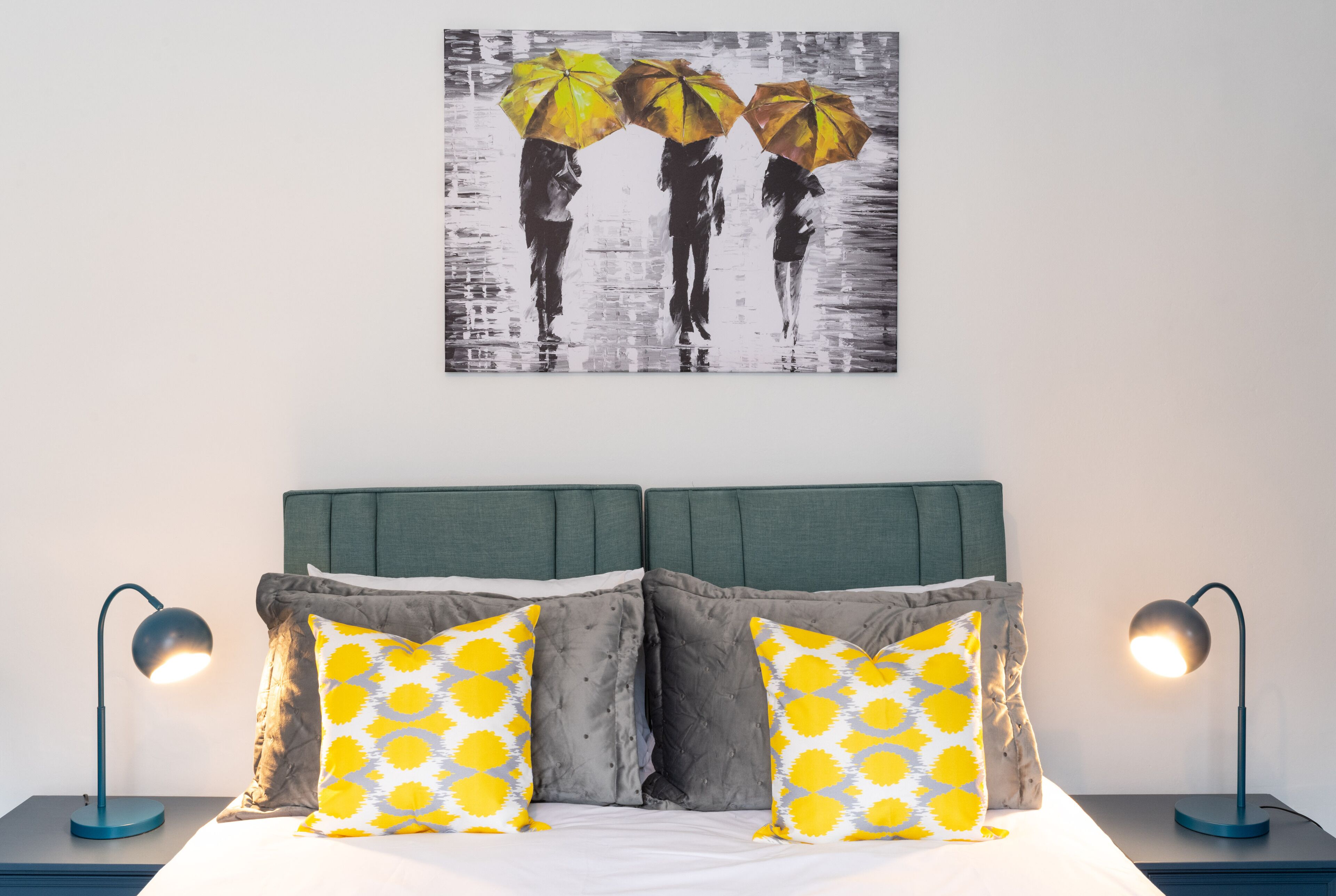 A neatly made bed with two pillows, a wall-mounted painting of three people with umbrellas, and two bedside lamps on either side.