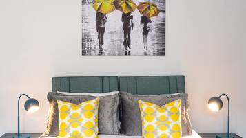 A neatly made bed with two pillows, a wall-mounted painting of three people with umbrellas, and two bedside lamps on either side.