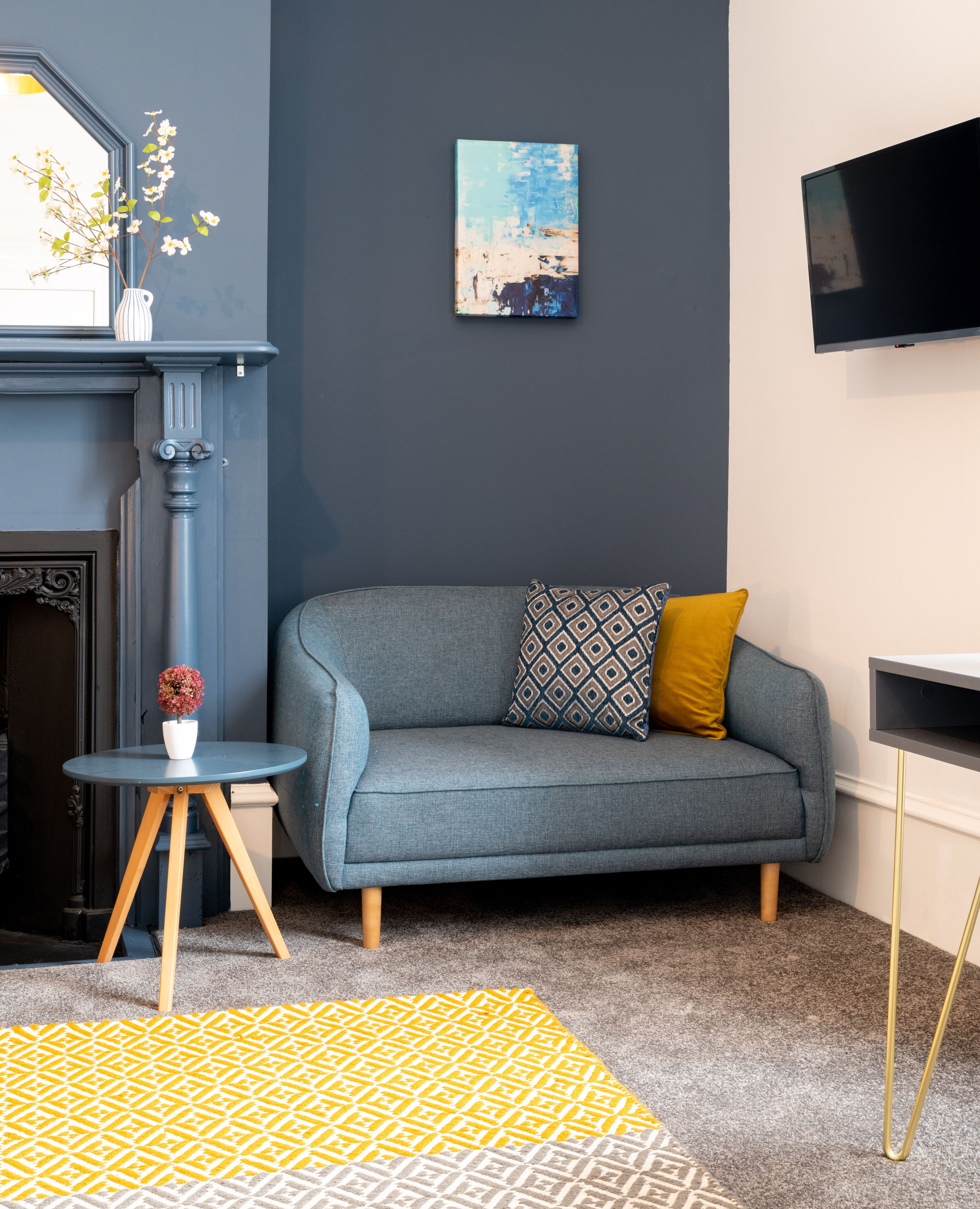 A modern living room with a grey sofa, a small round table with a vase, a patterned rug, and a fireplace.