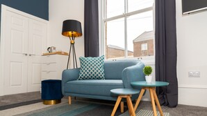 A modern living room with a blue sofa, a small round table, a floor lamp, and a potted plant. - Bricksage Rooms, King's Lynn South Gate (King's Lynn)