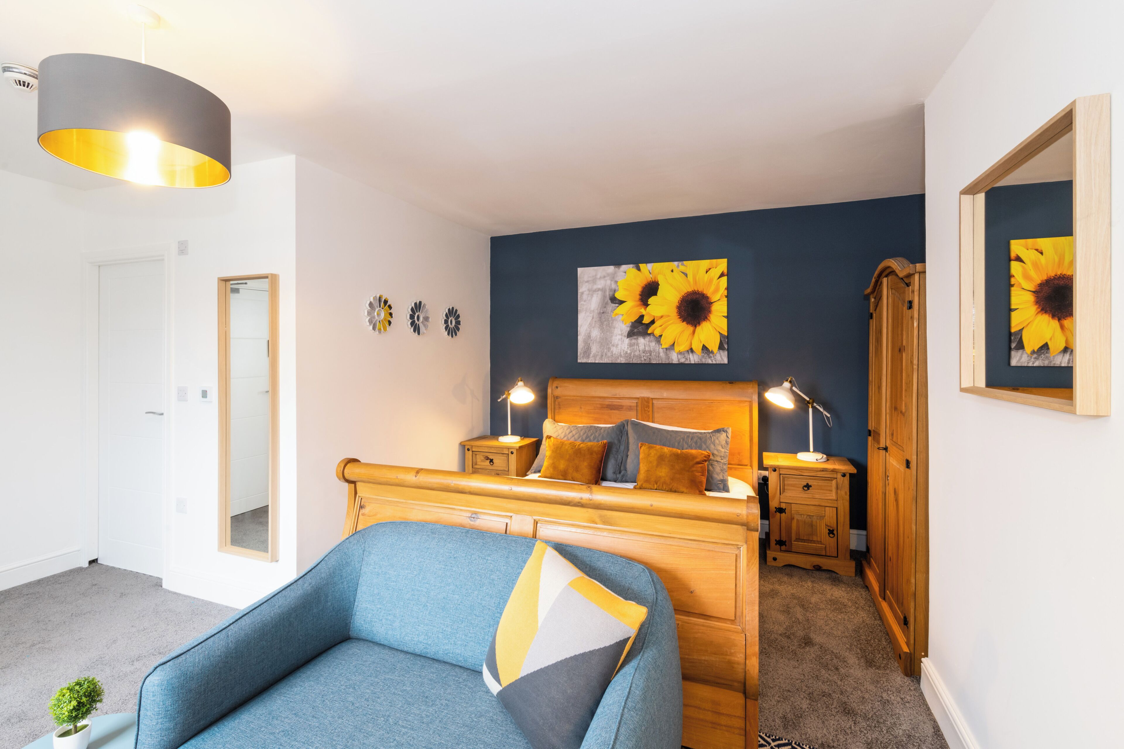 A bedroom with a bed, bedside tables, a lamp, a mirror, and a wall with a sunflower painting.