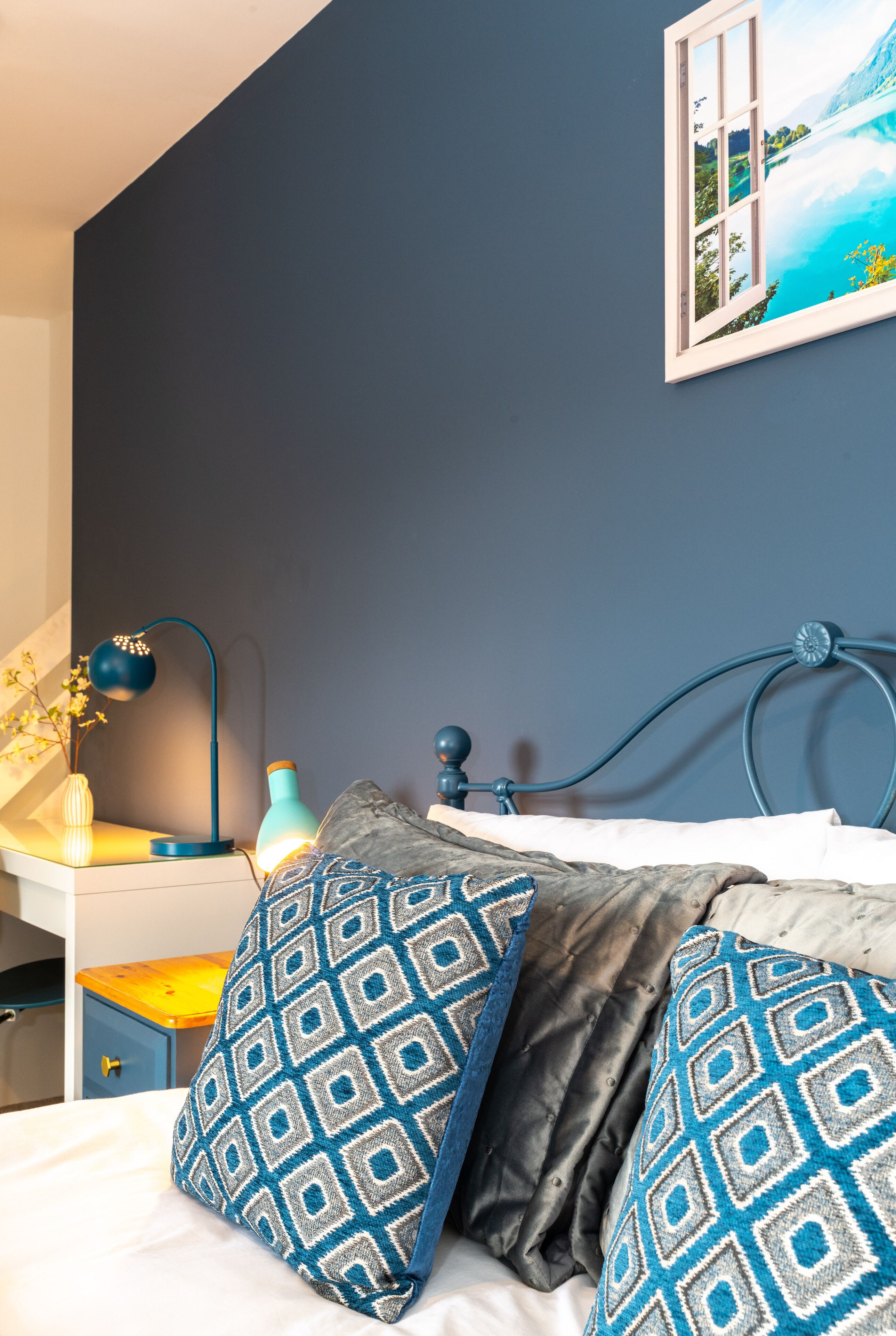 A bedroom with a bed, a nightstand, a lamp, and a wall painting.