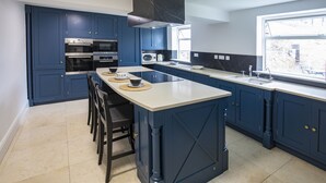 A modern kitchen with dark blue cabinets, a central island, and a large window providing natural light. - Bricksage Rooms, King's Lynn South Gate (King's Lynn)