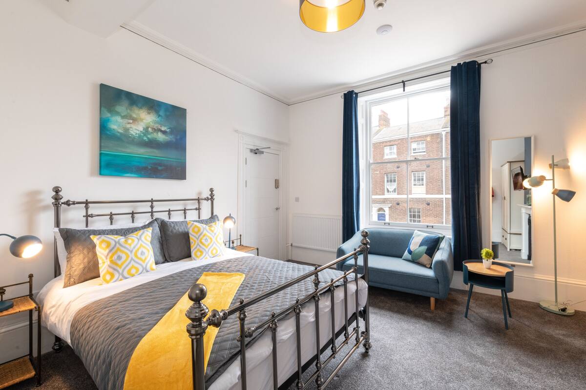A modern bedroom with a bed, a blue sofa, a window with curtains, a painting, and a mirror.