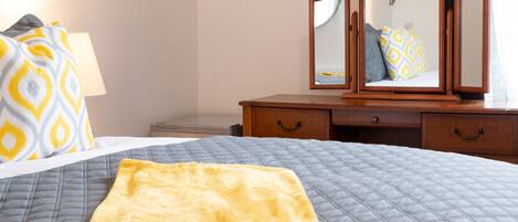 A neatly made bed with a quilted grey and blue cover, a yellow throw blanket, and a wooden dresser with two drawers and a mirror above it.
