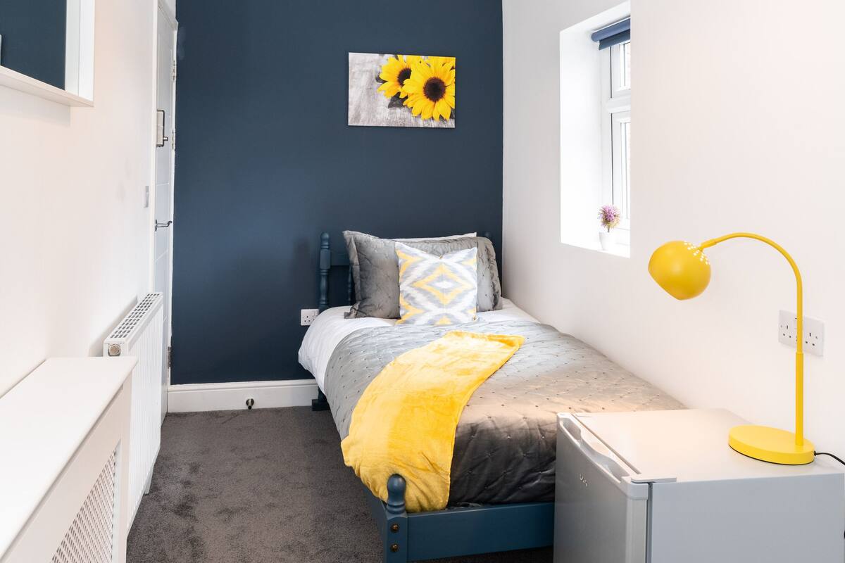 A compact bedroom with a bed, a bedside table, a lamp, a radiator, and a wall-mounted picture.