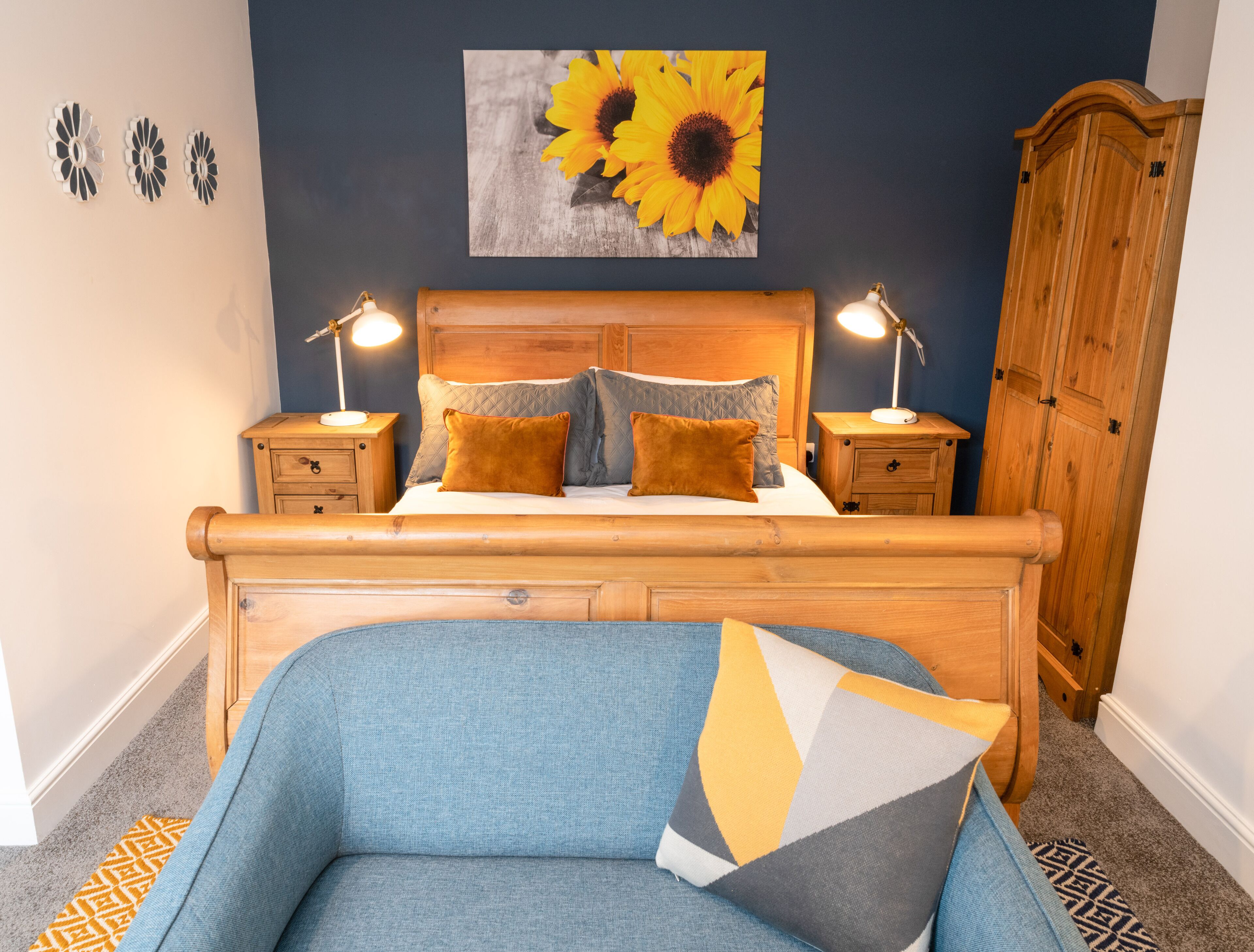 A bedroom with a wooden bed, two bedside tables with lamps, a large wooden wardrobe, and a wall with a sunflower painting.