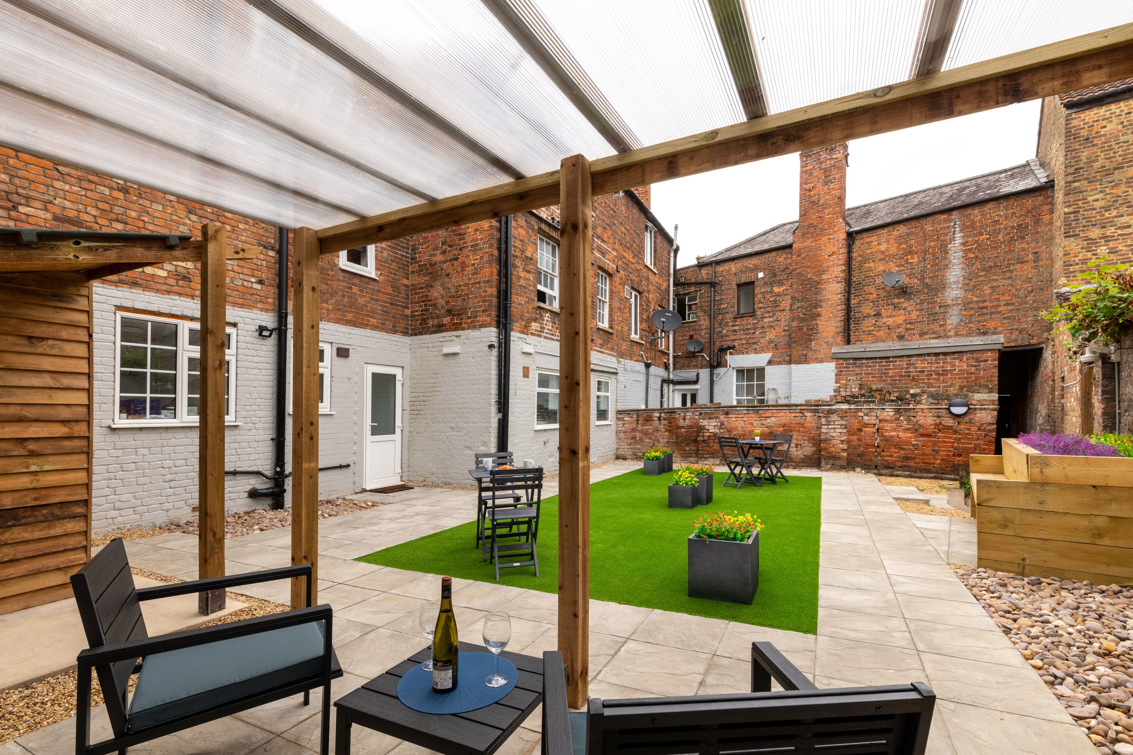 A courtyard with a wooden pergola, a patio table with a bottle and glass, and a seating area.