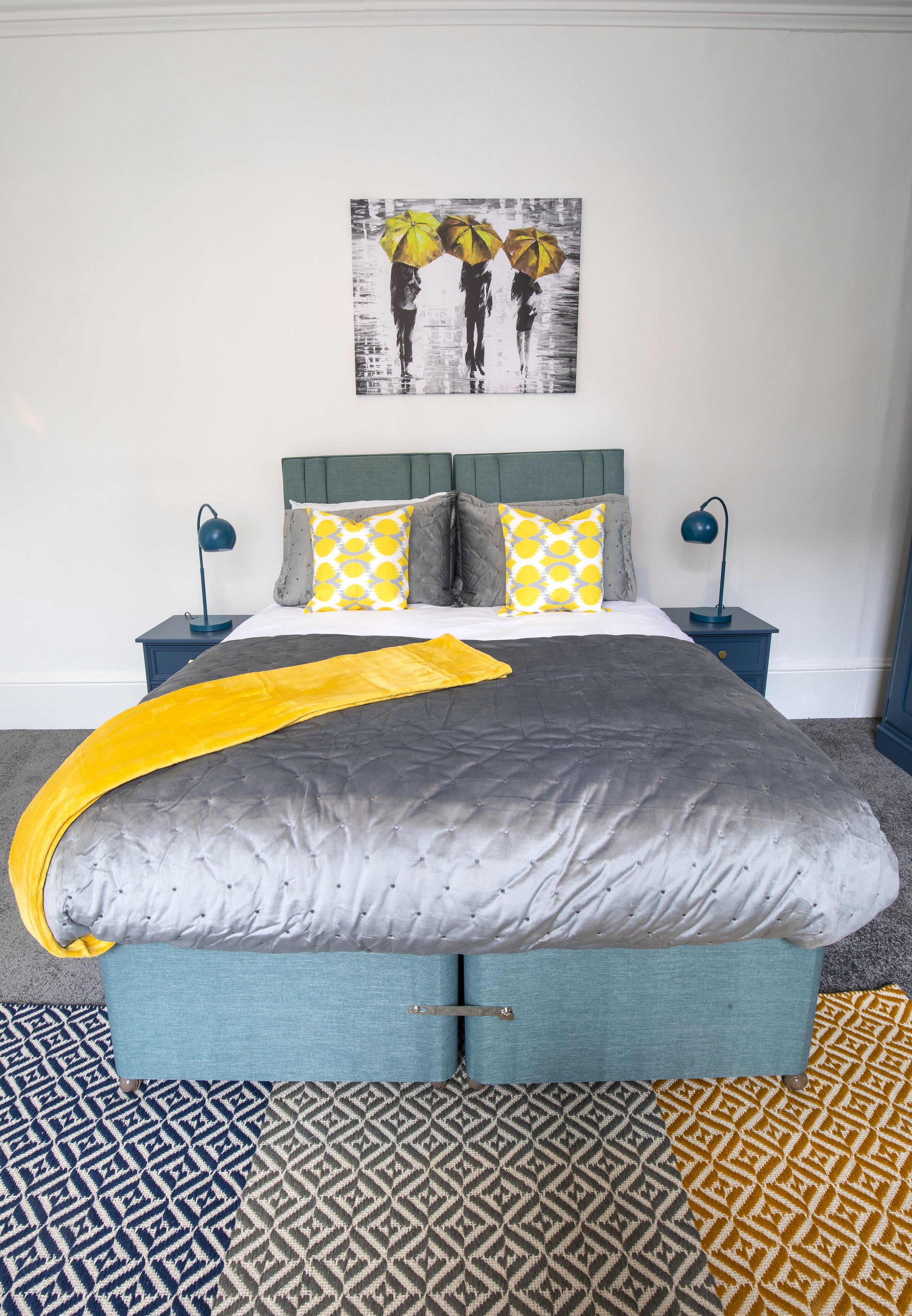 Un dormitorio con colcha gris, dos almohadas a juego, un plaid amarillo y una alfombra estampada.