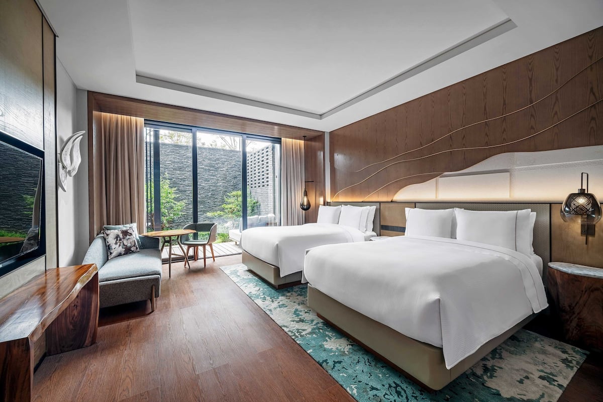 Een moderne hotelkamer met twee bedden, een zithoek en grote ramen met uitzicht op groen.