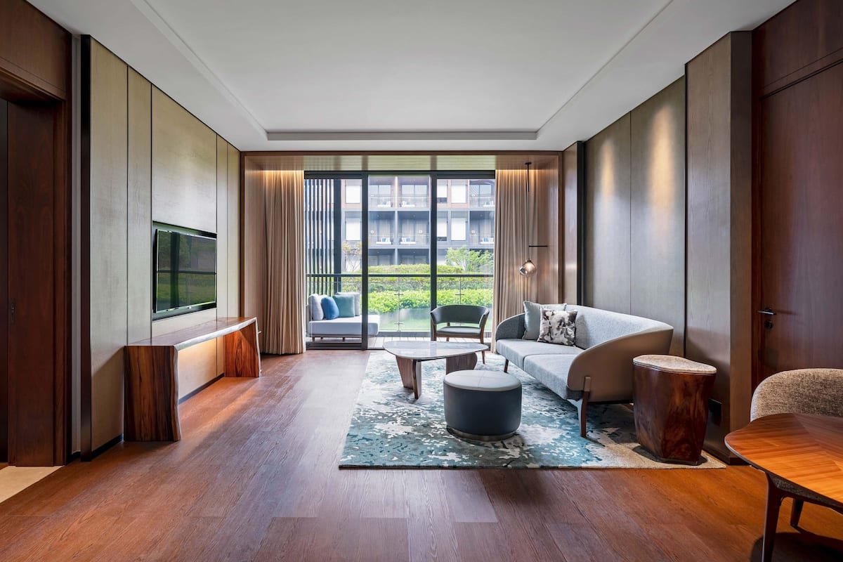 Een moderne woonkamer met een bank, salontafel en voetenbankje, een flatscreen-tv en een groot raam met uitzicht op een gebouw en groen.