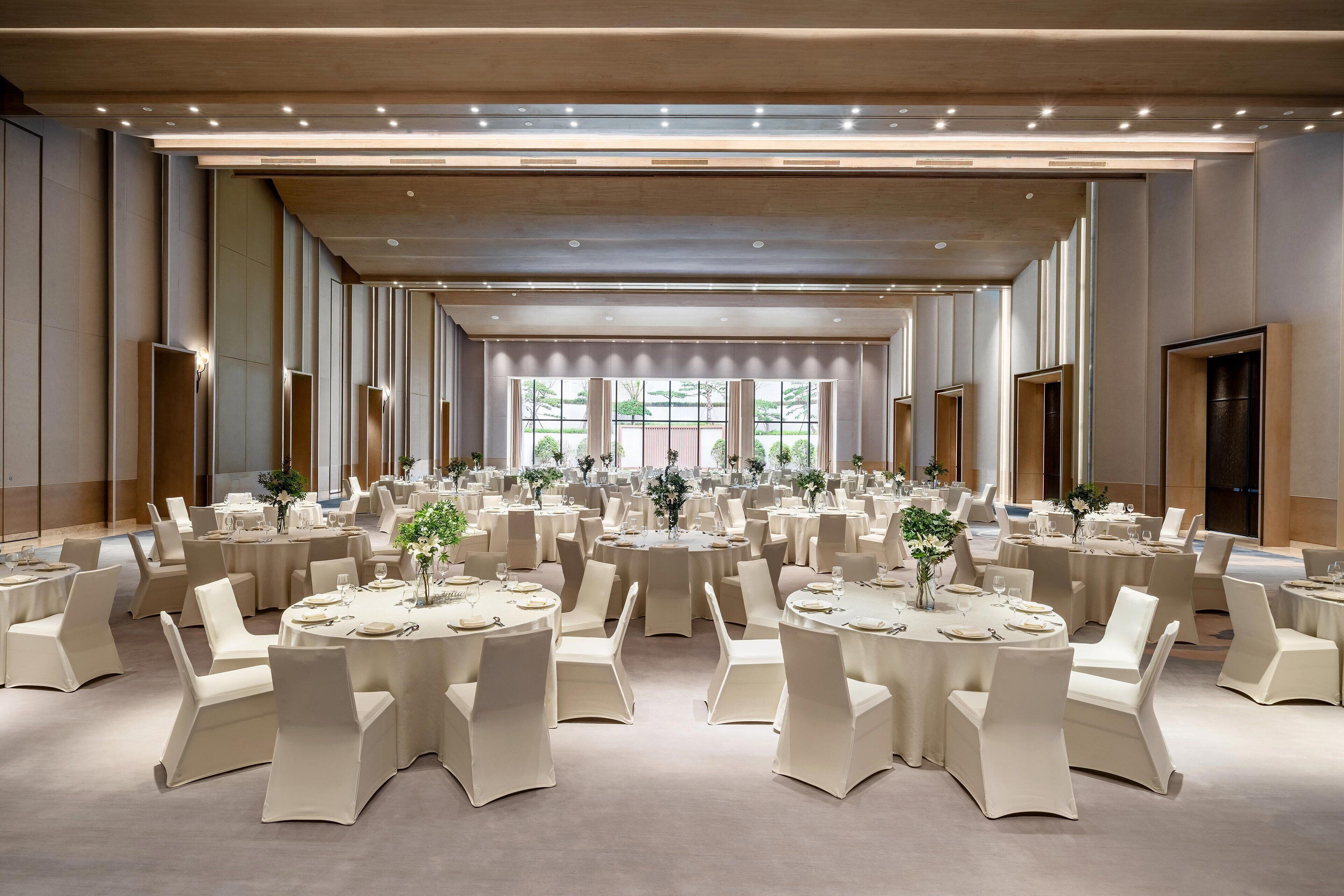 Un amplio salón de banquetes con varias mesas redondas, cada una con manteles blancos y sillas, y grandes ventanales que aportan luz natural.