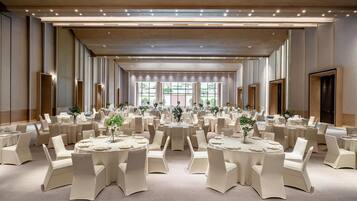 Un amplio salón de banquetes con varias mesas redondas, cada una con manteles blancos y sillas, y grandes ventanales que aportan luz natural.