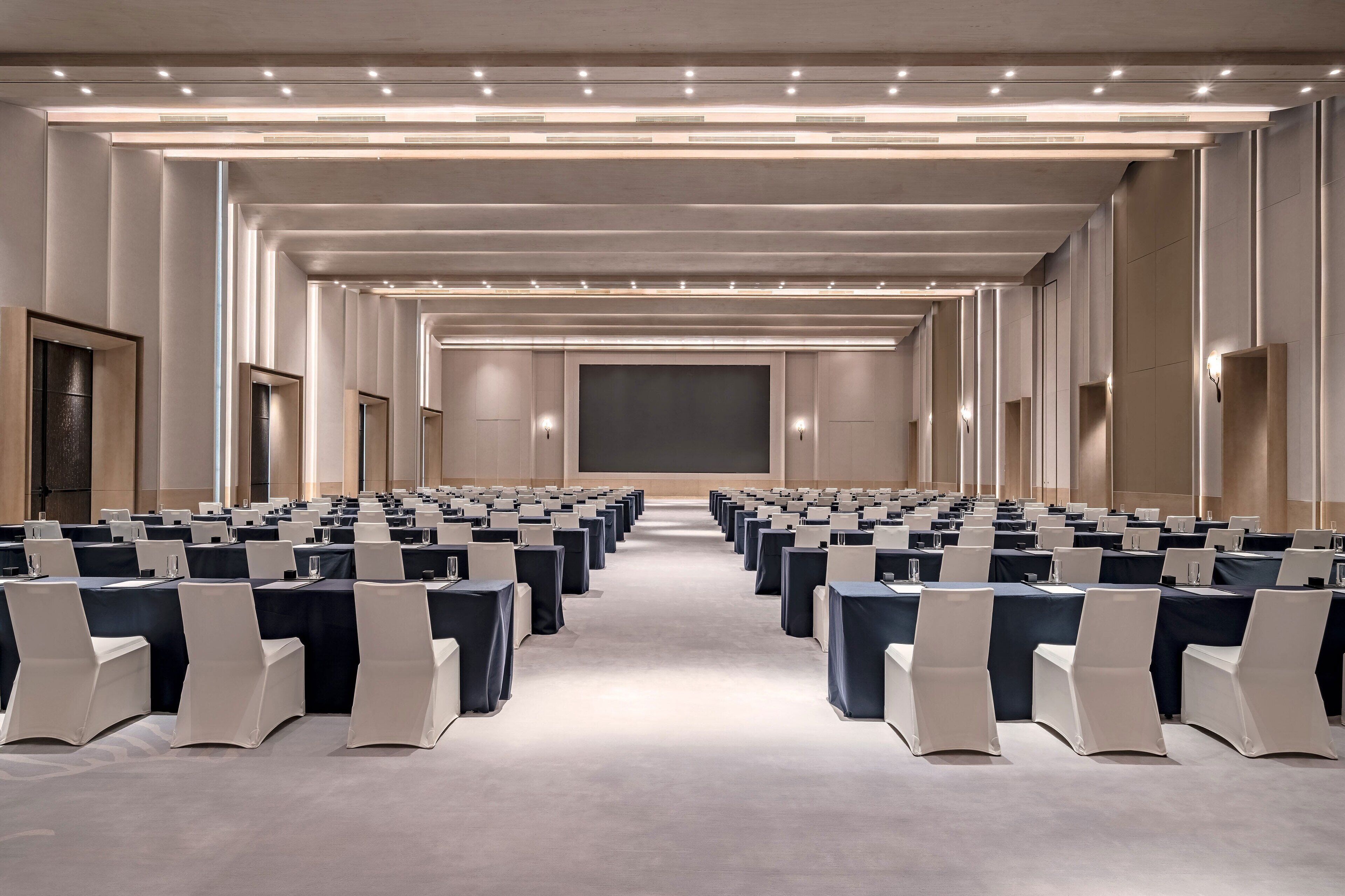 Une salle de conférence avec des rangées de tables et de chaises, un grand écran à l’avant et un éclairage encastré.