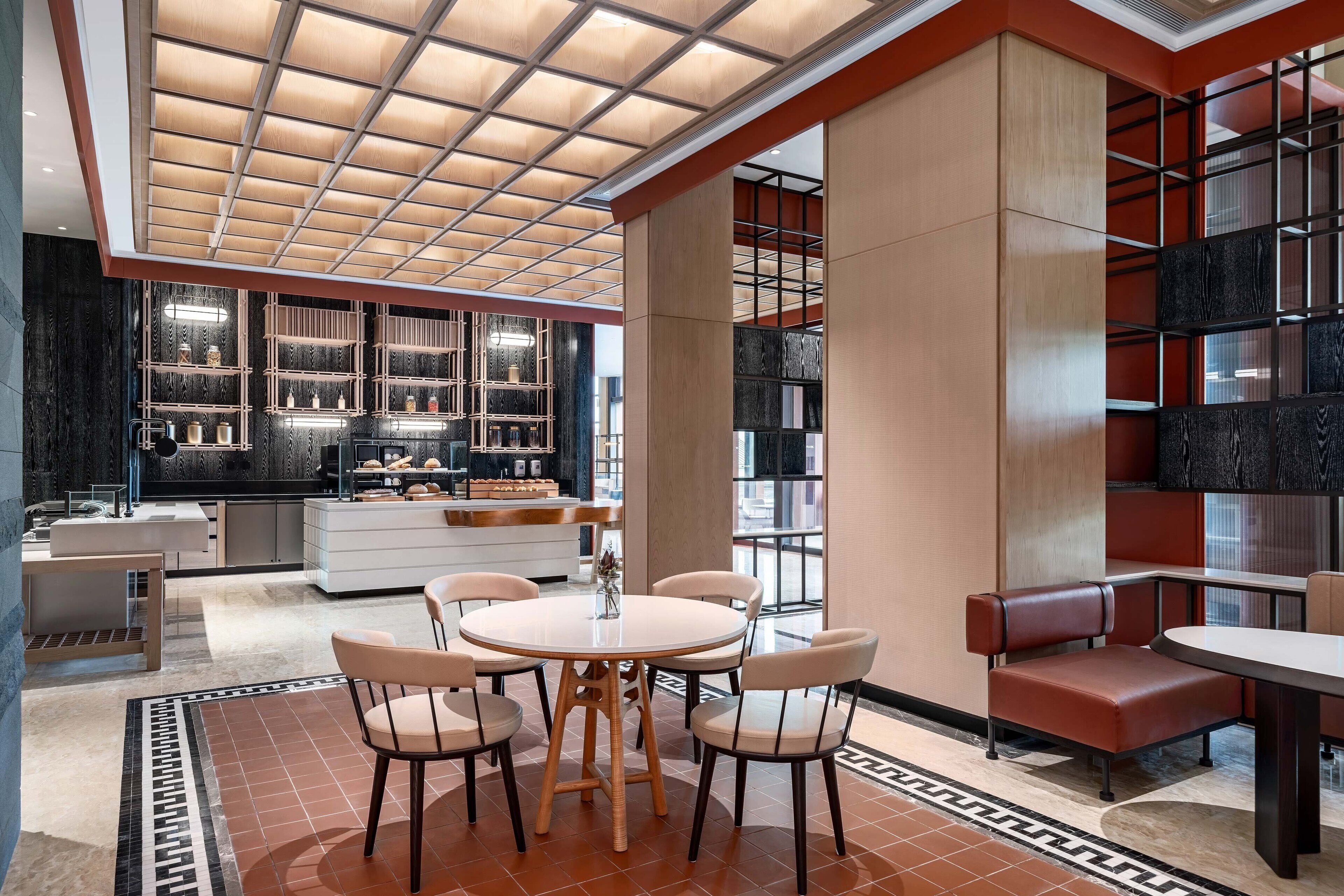A modern hotel lobby with a central round table, seating area, and a kitchen in the background.
