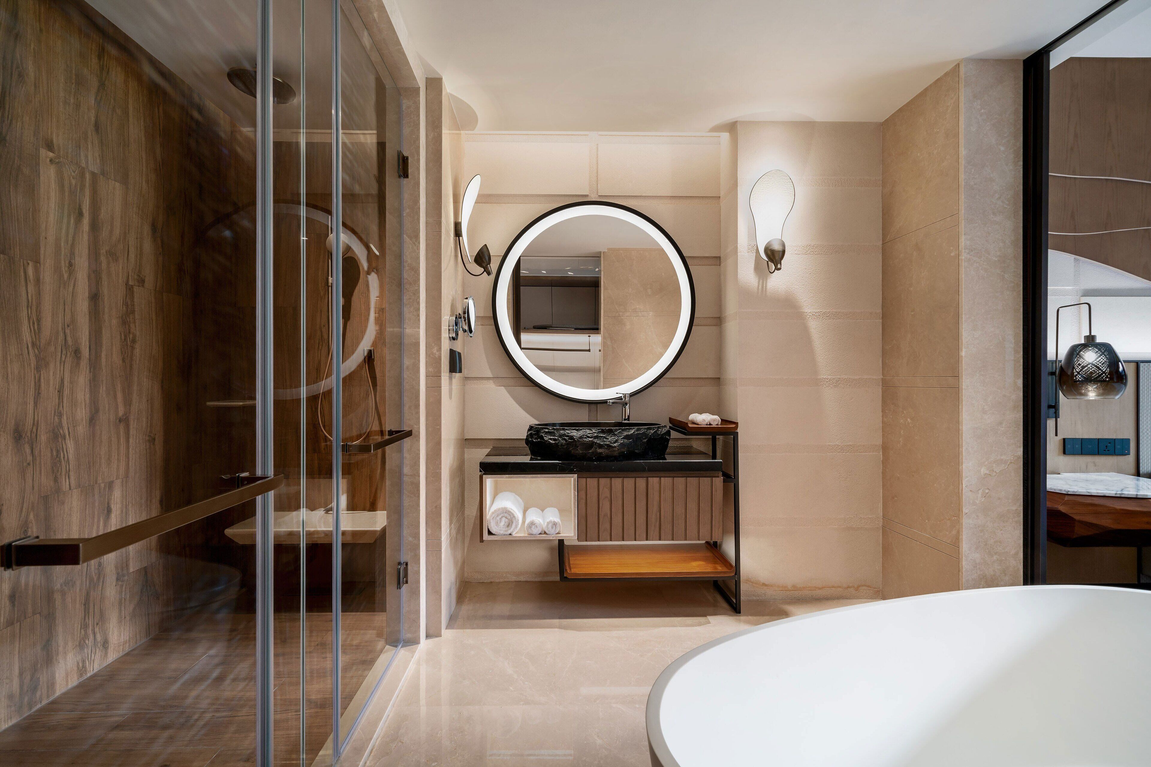 A modern bathroom with a glass shower enclosure, a round mirror with a light, a sink with a black basin, and a wooden vanity.