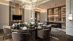 Un espace de salle à manger moderne, avec une table ronde pour six personnes, entourée de chaises rembourrées. Un téléviseur est fixé au mur, et des étagères ornées d’objets décoratifs se trouvent à l’arrière-plan.