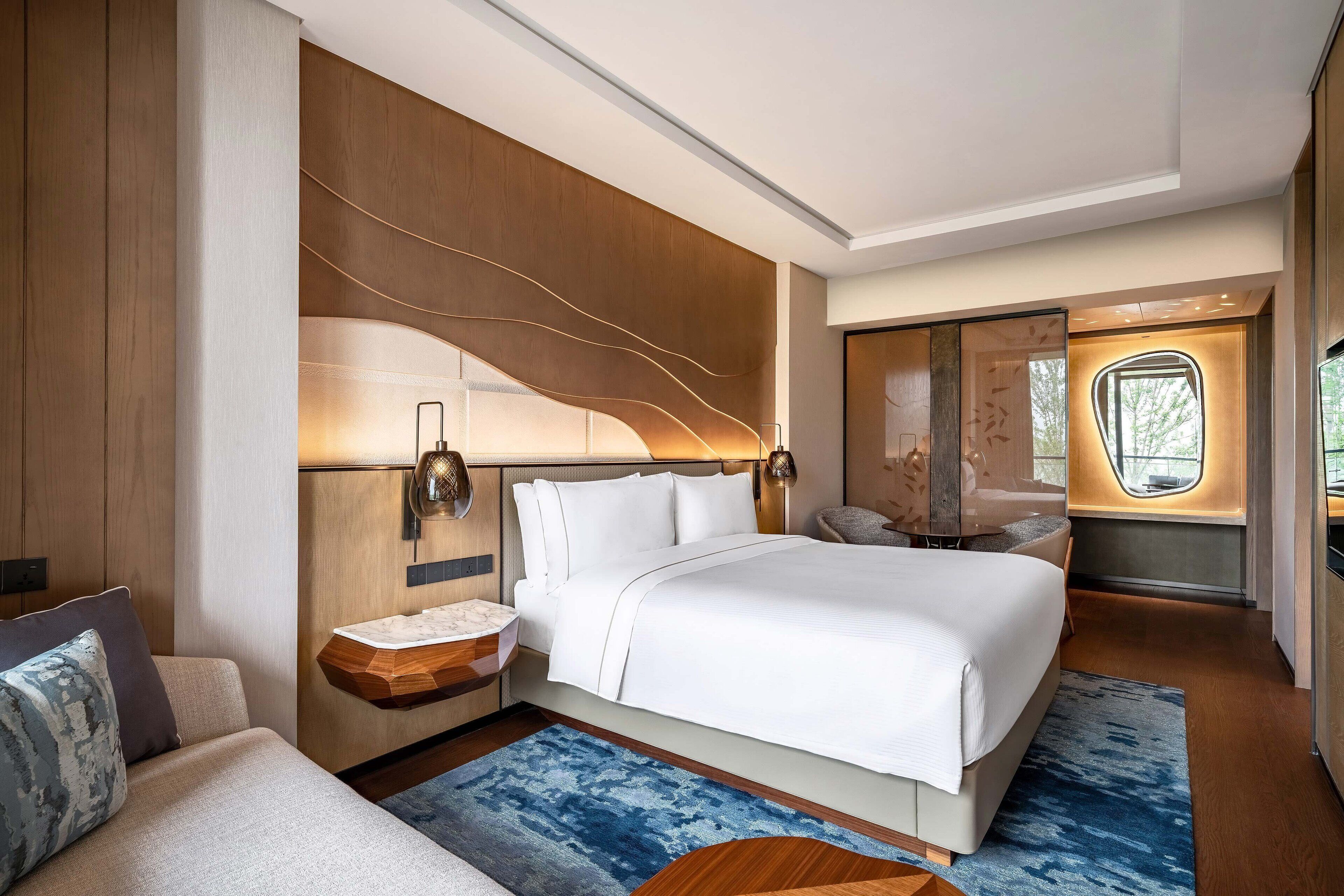 Ein modernes Hotelzimmer mit einem großen Bett, einem Nachttisch, einer integrierten Sitzbank und Blick ins Freie.