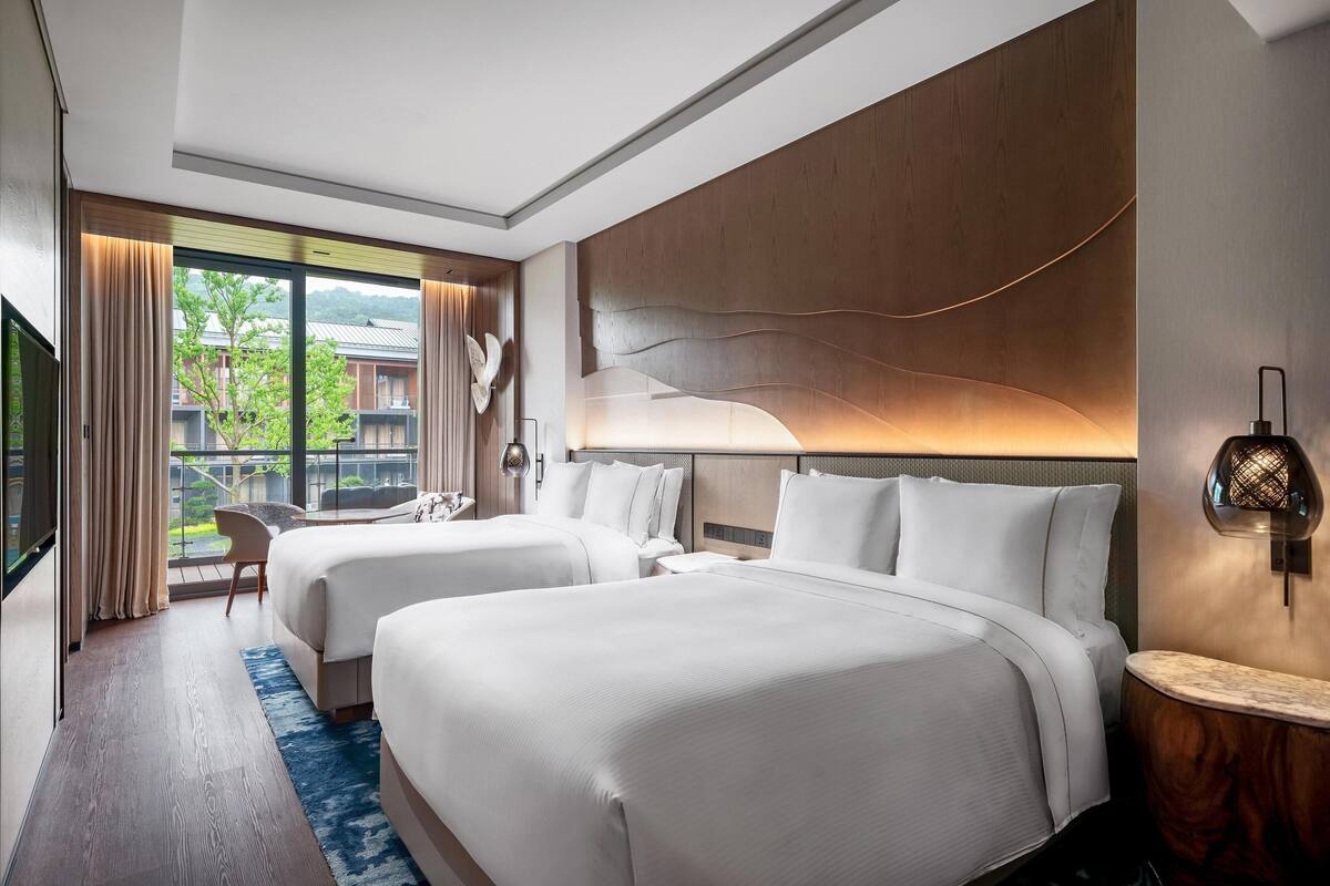 Een hotelkamer met twee bedden, een groot raam met gordijnen, een houten hoofdeinde en een nachtkastje met een lamp.