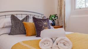 Una cama bien tendida con cabecera de metal, un plaid amarillo y blanco estampado, y toallas enrolladas.