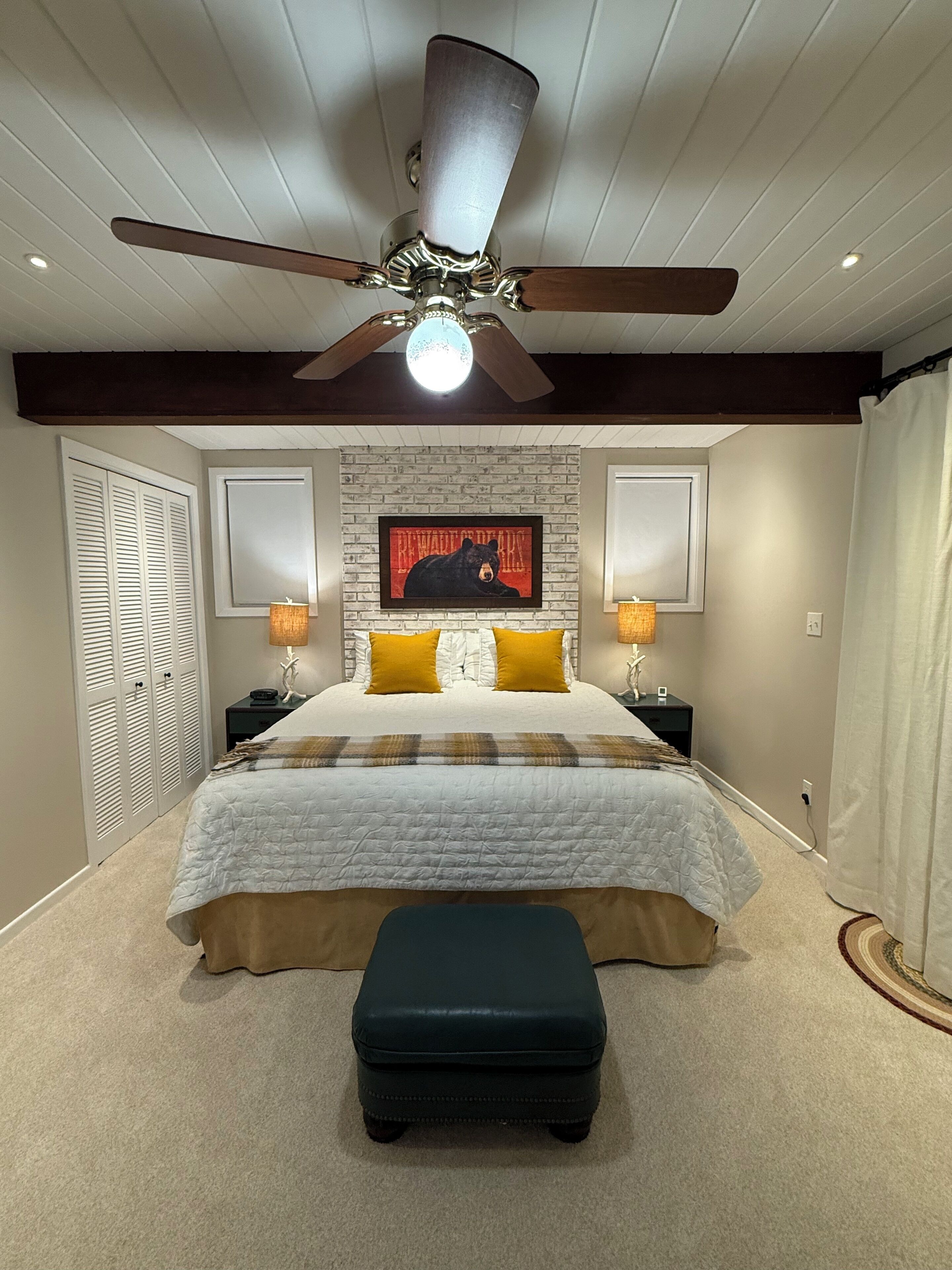 A bedroom with a bed, a ceiling fan, a wall-mounted painting, bedside lamps, and a bench.
